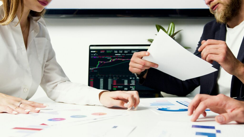 Two people review investment account statements. There is a computer displaying a stock ticker in the background. 