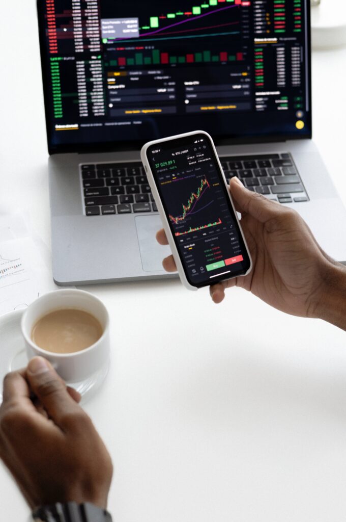 Hand holding a smartphone with stock ticker in front of a laptop that also displays a stock portfolio. The other hand holds a cup of coffee. 